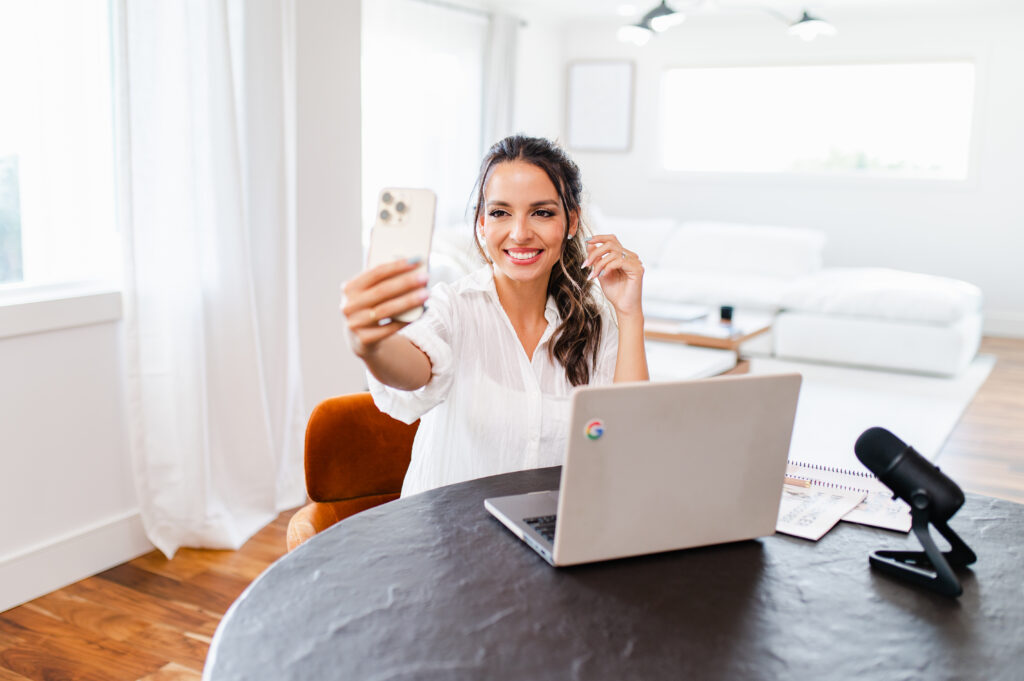 Influencer smiling while recording an Instagram Story with her smartphone, sitting in a modern workspace with a laptop and microphone, showcasing how to improve Instagram story quality.